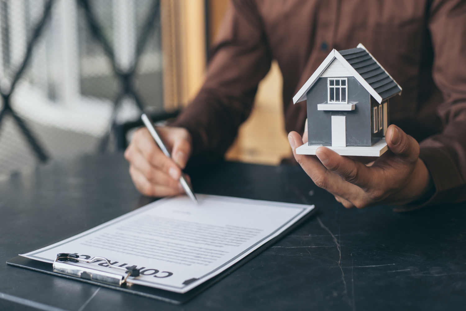 Pessoa segurando uma miniatura de casa enquanto assina um contrato em uma prancheta, representando um acordo imobiliário ou transação de compra e venda de imóveis.